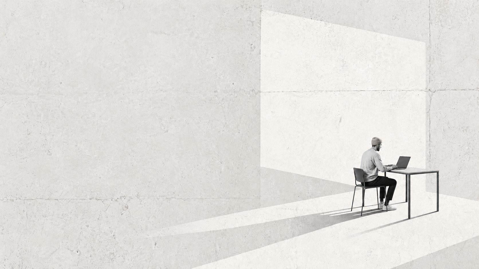A man in an ochre beanie working at a laptop in a sunlit concrete room